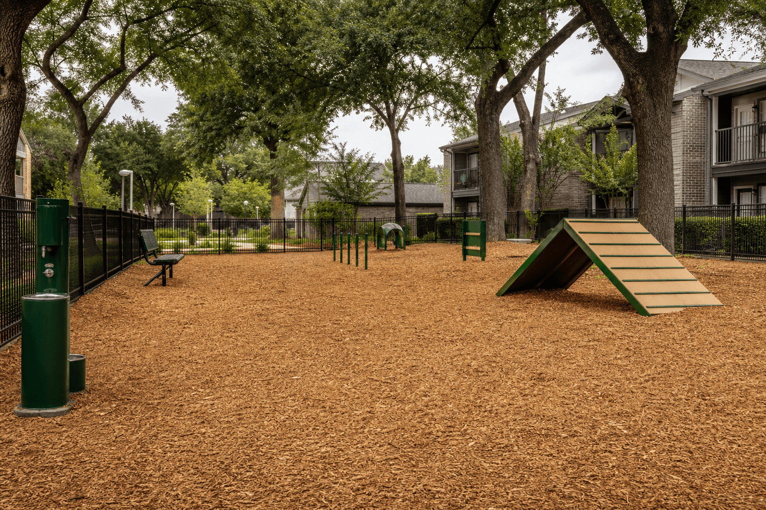 A fenced apartment dog park with a freshly spread wood chip run, shade trees and agility equipment
